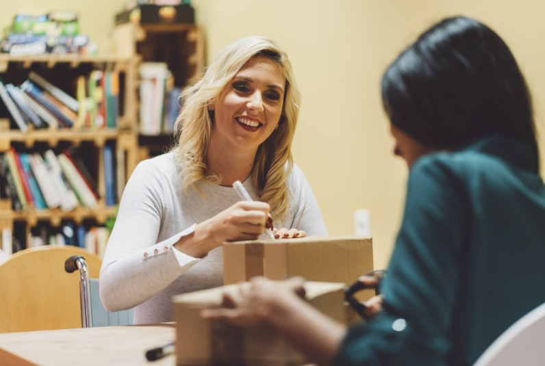 Two women sit at a table in a room with bookshelves, smiling as they write on cardboard boxes.