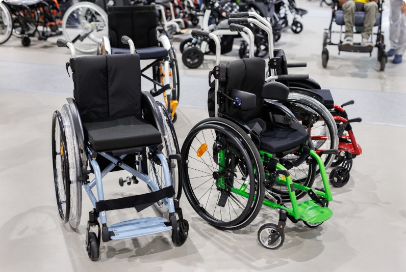 Several wheelchairs of different designs and colours are displayed indoors on a light-coloured floor, with more wheelchairs visible in the background.