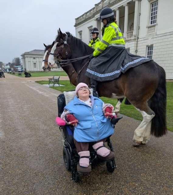A woman in a wheelchair sits on a path whilst two mounted police officers on horses are nearby in front of a large white building.
