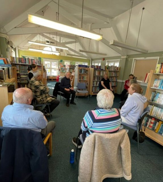 Eight adults sit in a circle having a discussion in a library surrounded by bookcases and natural light.