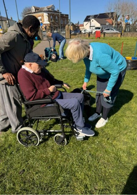 An older man sits in a wheelchair on grass, accompanied by two adults; one stands behind him, the other leans forward holding a lead. A black and white dog is next to them.