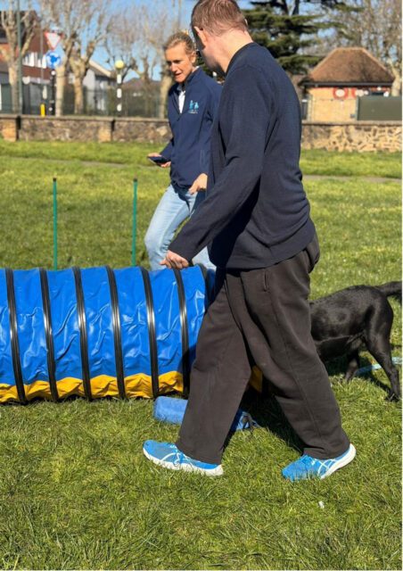 A man and a woman stand near a blue and yellow dog agility tunnel on grass, with a black dog entering the tunnel.