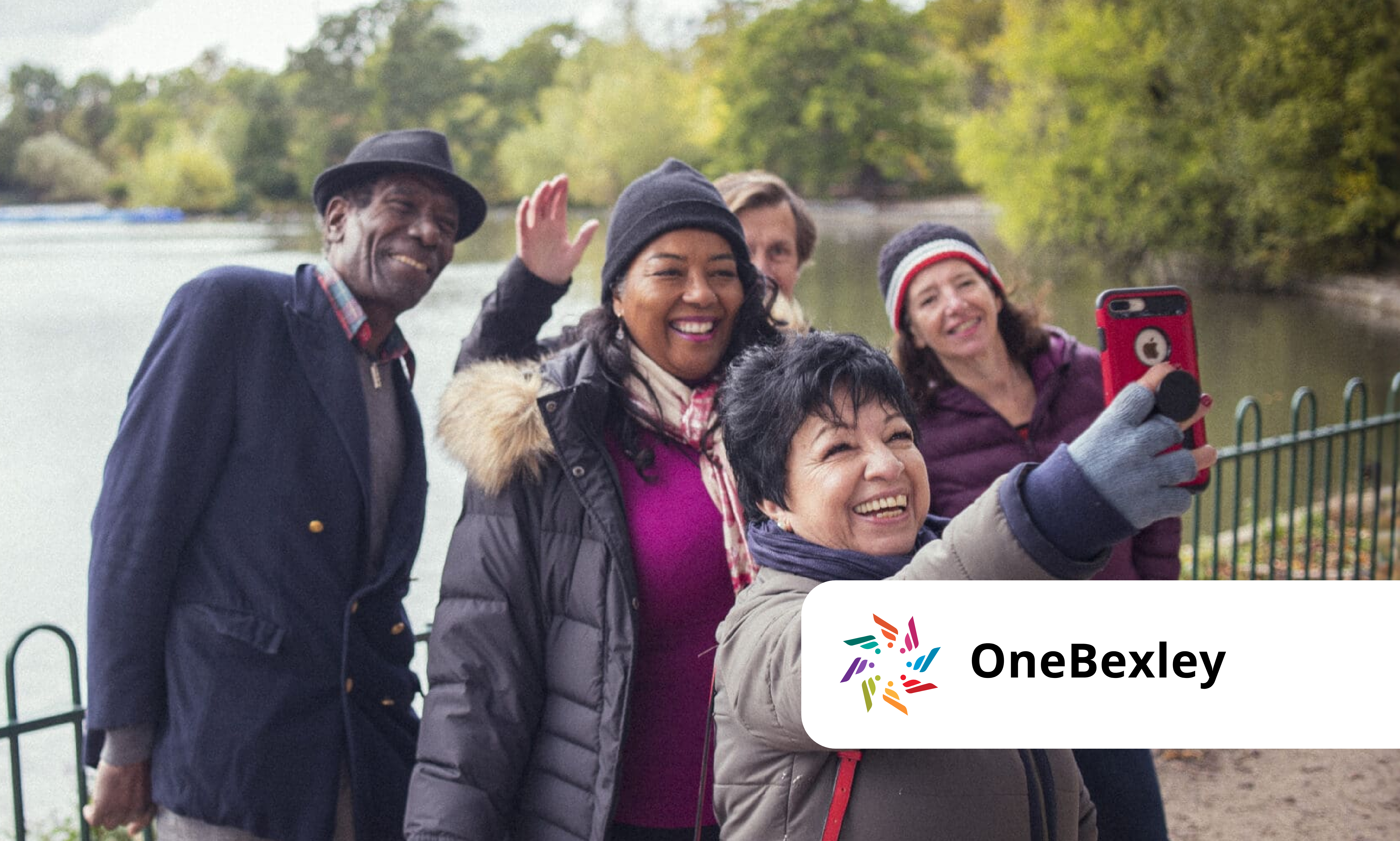 A group of five adults in warm clothing smile and take a selfie by a riverside path, with trees in the background. The OneBexley logo is displayed in the bottom right corner.